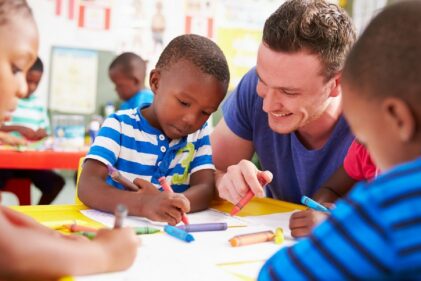An early childhood education teacher looks on as his young students draw with crayons.