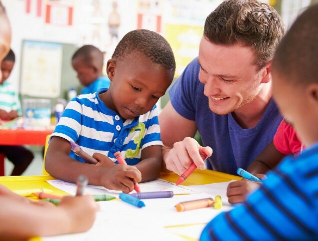 An early childhood education teacher looks on as his young students draw with crayons.