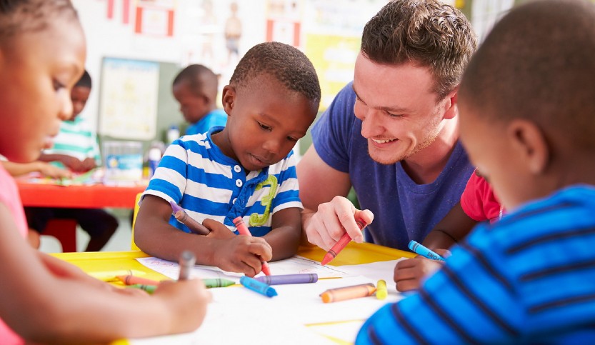 An early childhood education teacher looks on as his young students draw with crayons.