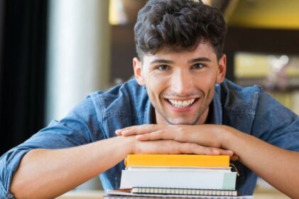 A man smiles at the camera while resting his hands on a stack of books and his chin on his hands.