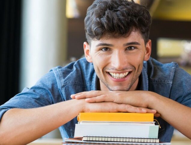 A man smiles at the camera while resting his hands on a stack of books and his chin on his hands.