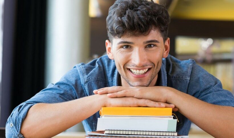 A man smiles at the camera while resting his hands on a stack of books and his chin on his hands.