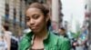A young women wearing a green jacket stands on a city street and looks directly at the camera with a neutral expression.
