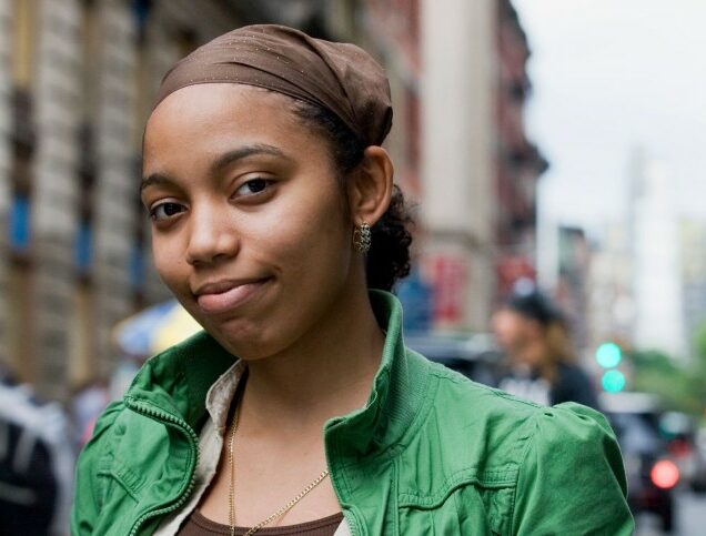 A young women wearing a green jacket stands on a city street and looks directly at the camera with a neutral expression.