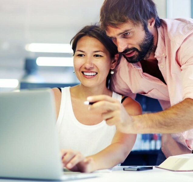 A woman seated at a desk looks at a laptop computer as a man leaning over her shoulder points at something on the screen.