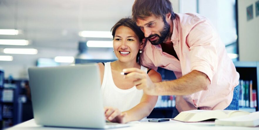 A woman seated at a desk looks at a laptop computer as a man leaning over her shoulder points at something on the screen.