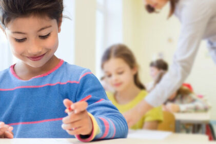 A young elementary school student smiles and holds a writing utensil at their desk while an instructor instructs another student in the background.