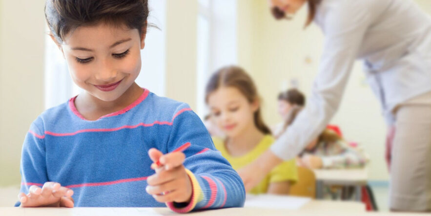 A young elementary school student smiles and holds a writing utensil at their desk while an instructor instructs another student in the background.