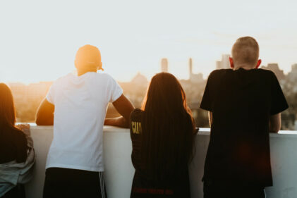 Four young adults stand on a rooftop, watching a sunrise with their backs to the camera.