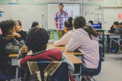 A group of children in a classroom pay attention to a man standing before them at the front of the room.
