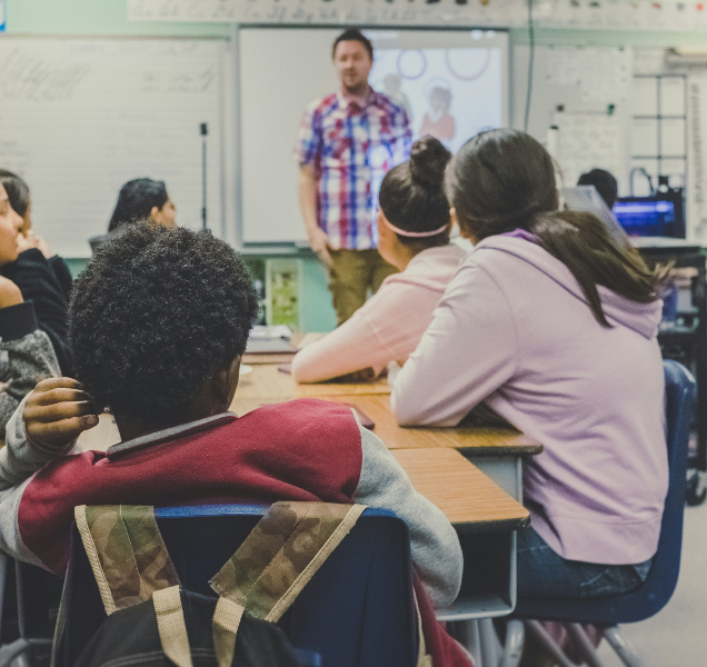 A group of children in a classroom pay attention to a man standing before them at the front of the room.
