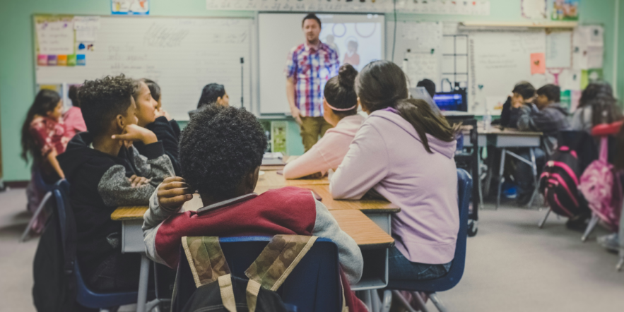 A group of children in a classroom pay attention to a man standing before them at the front of the room.