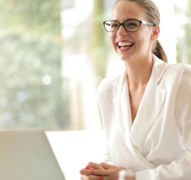 A smiling person wears glasses and a white blouse while seating at a desk with a laptop in front of them.