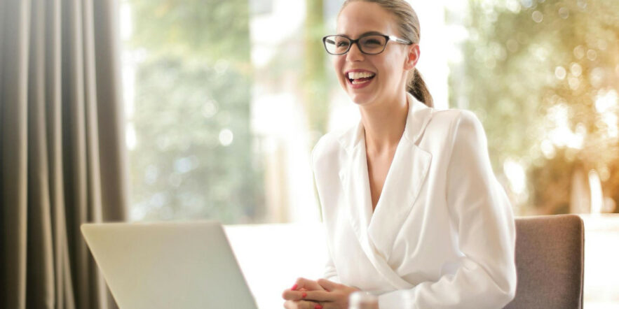A smiling person wears glasses and a white blouse while seating at a desk with a laptop in front of them.