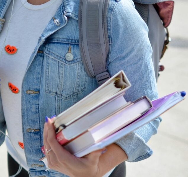 A student wearing a denim jacket carries books, binders and a backpack.