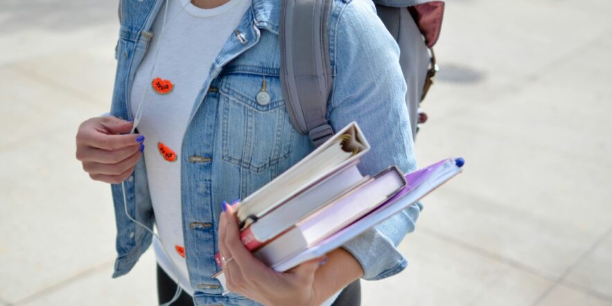 A student wearing a denim jacket carries books, binders and a backpack.