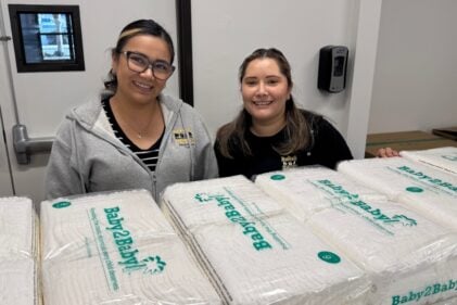Two people smile for the camera while behind packages of Baby2Baby diapers.