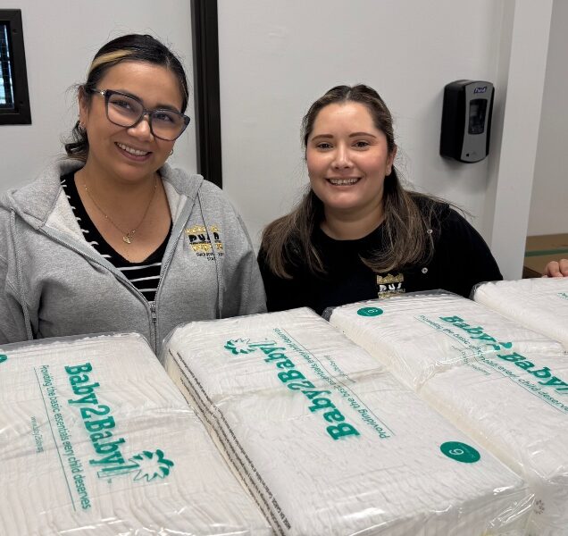 Two people smile for the camera while behind packages of Baby2Baby diapers.