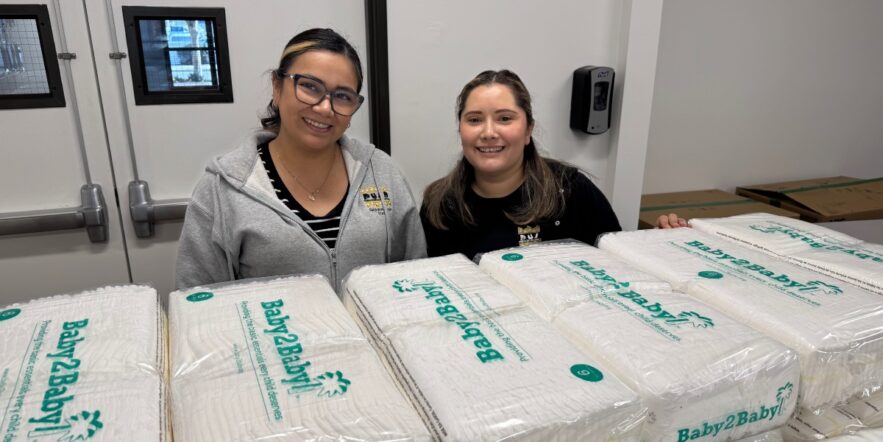 Two people smile for the camera while behind packages of Baby2Baby diapers.