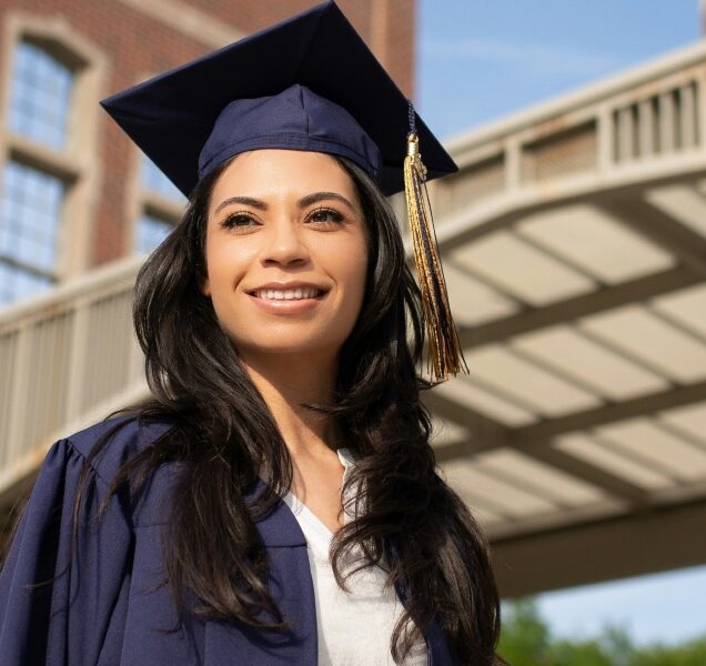 A person wears a graduate cap and gown and half-smile looks into the middle distance contemplating her life choices.