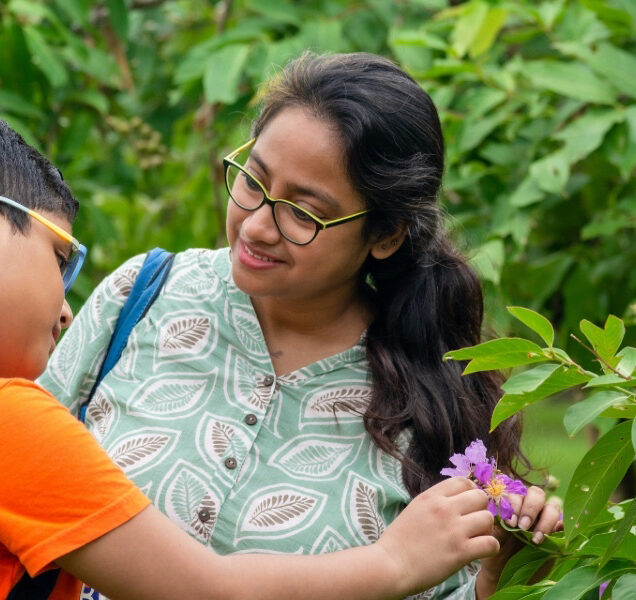 Woman shows flower to young child with backpack.