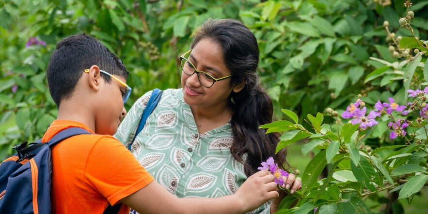 Woman shows flower to young child with backpack.