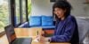 Woman with glasses sitting at a desk with a laptop, writing on a notepad.