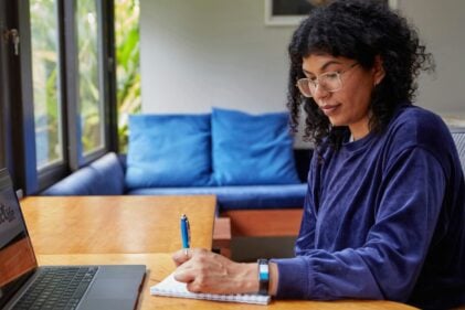 Woman with glasses sitting at a desk with a laptop, writing on a notepad.