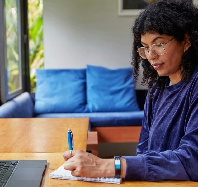 Woman with glasses sitting at a desk with a laptop, writing on a notepad.